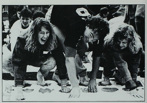Black and white image of Iowa State Students playing Twister. There are three students in the foreground, they look like they are laughing while posed in awkward positions on a Twister mat.