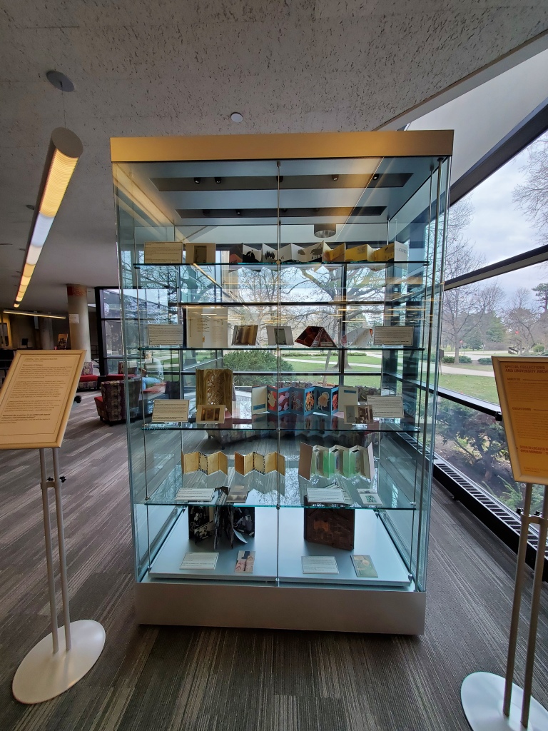 Glass exhibit case with five shelves on which are arranged several books